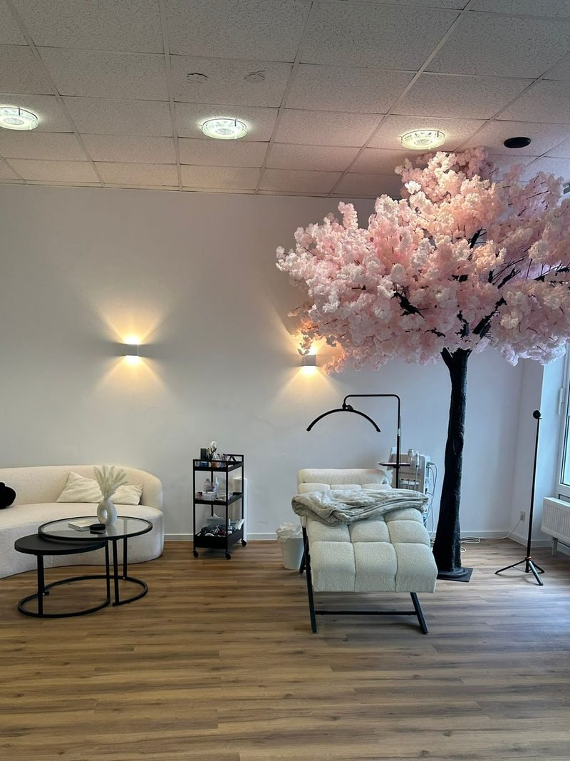 Behandlungsliege im Studio, dar&uuml;ber ein gro&szlig;er Kirschbl&uuml;tenbaum, Boucl&eacute;-Sofa und warme Wandleuchten