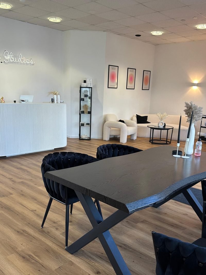 Blick in das Studio &mdash; Neon-Schriftzug &bdquo;Adorable Studios&ldquo;, wei&szlig;er Empfangstresen, Lounge-Bereich mit gerahmten Bildern und gro&szlig;er Arbeitstisch im Vordergrund